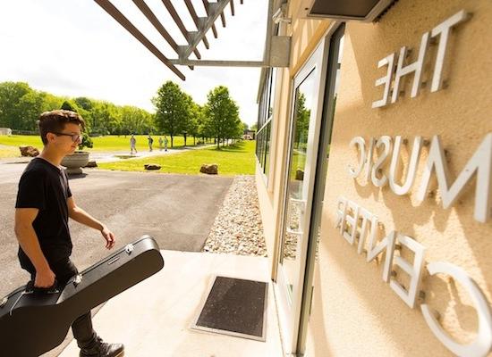 A young person with short hair and glasses, wearing a black outfit, walks by "THE MUSIC CENTER" sign on a sunny day, carrying a guitar case, indicating they are possibly heading to or from a music lesson or performance.